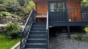 Une cabane en bois avec un escalier menant à une terrasse, entourée d’arbres et d’un mur de pierres.