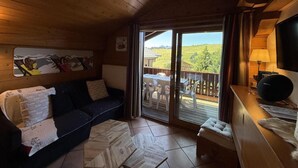 Een gezellige kamer met een houten plafond, een bank, een klein tafeltje en uitzicht op een balkon met tuinmeubilair.