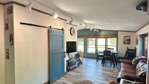 A spacious living area with a large brown leather sofa, a dining table with chairs, and a TV mounted on the wall. - Livingston MT Paradise Unit B (Livingston)