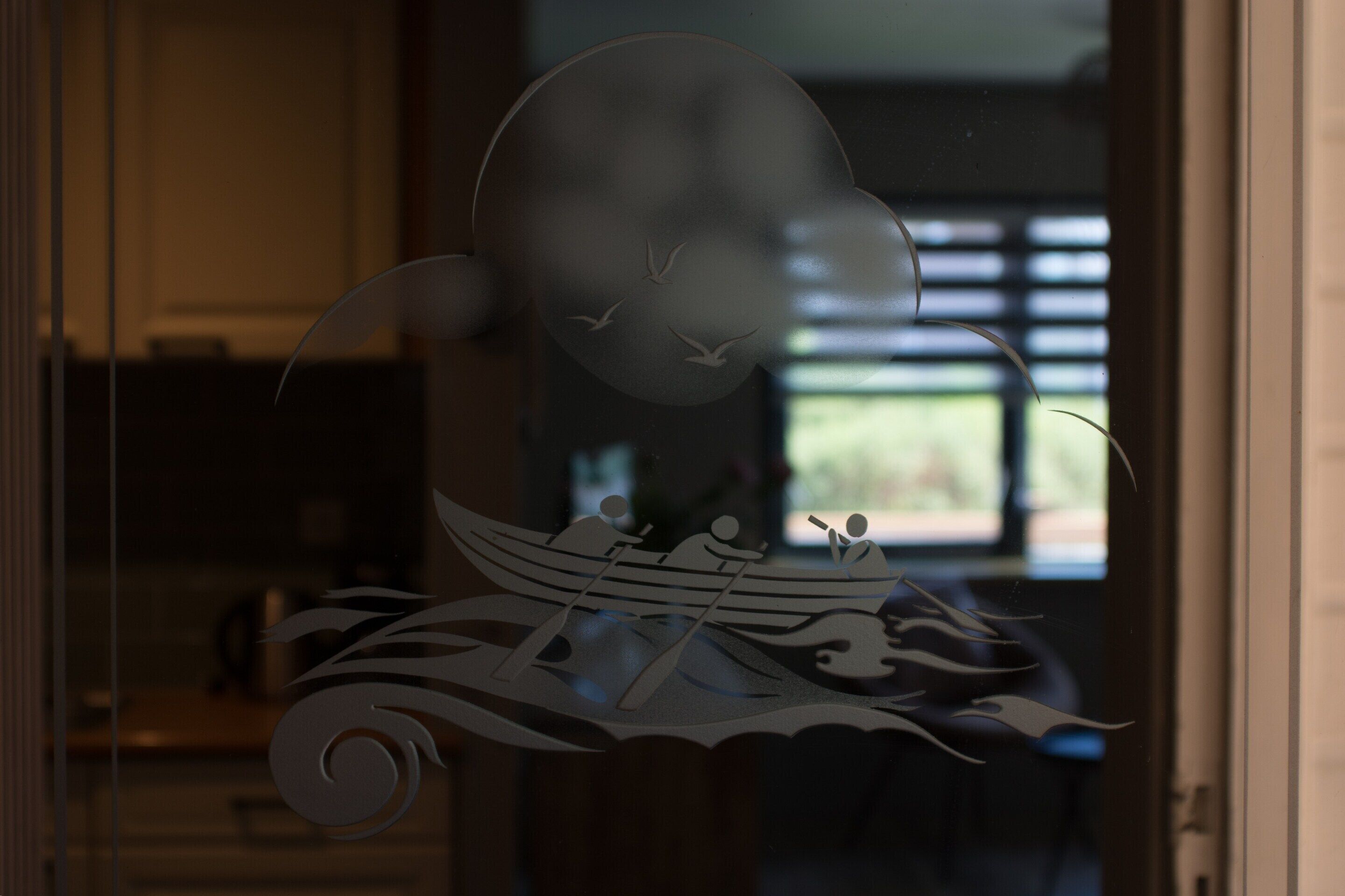 Een decoratief glazen raam met een silhouet van een boot en figuren, mogelijk een uitzicht op een waterlichaam.