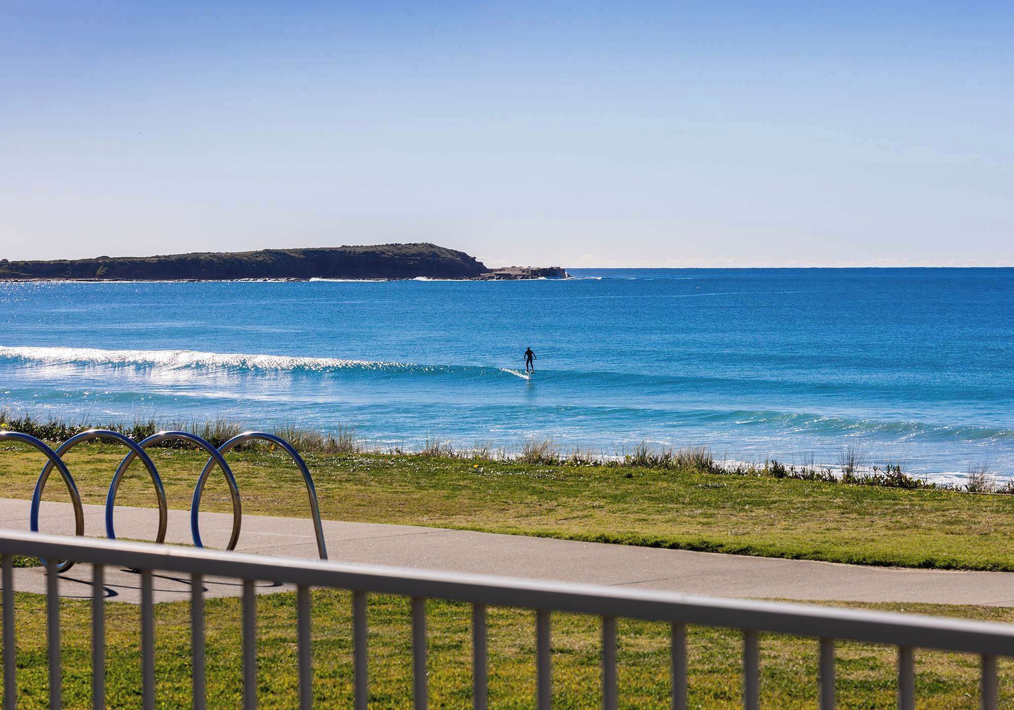 A coastal view with a person surfing, a grassy area, and a metal railing.