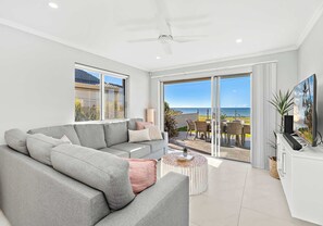 Un salon moderne avec un canapé gris, une petite table basse ronde et un téléviseur posé sur un meuble blanc. De grandes fenêtres offrent une vue sur l’océan et sur le mobilier d’extérieur installé sur la terrasse.