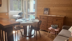 A cozy cabin interior with a wooden dining table set for four, surrounded by chairs. A small round table with a blue bowl is visible in the foreground.
