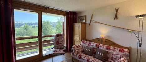 A cozy room with a patterned sofa, a wooden table, and a view through large sliding glass doors to a balcony.