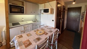 A compact kitchen with a microwave, stove, and sink. There is a dining table with a quilted tablecloth and a decorative plate. The kitchen has wooden cabinets and a wooden ceiling. - 100m from ski slopes, 2nd floor, view ski slopes, terrace, tv, ski locker, 24m², Les Saisies (Les Saisies)
