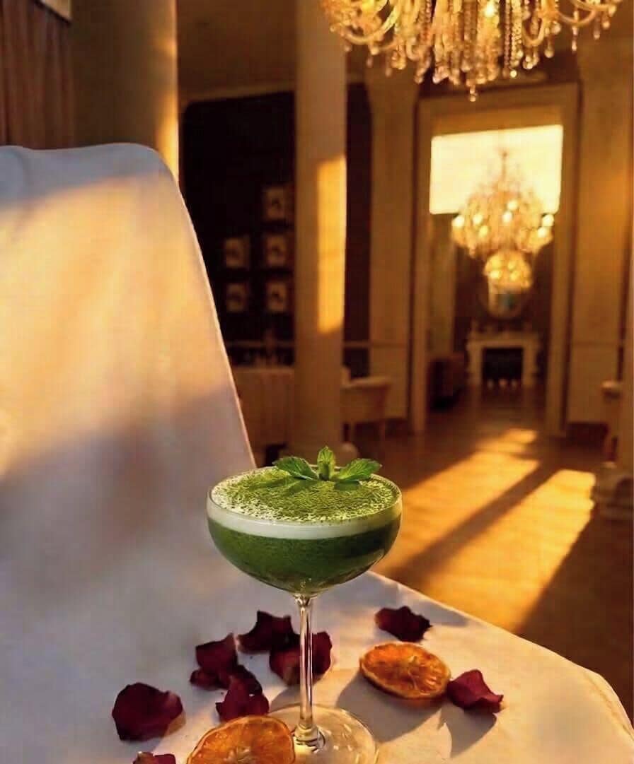A cocktail on a table with rose petals, in a room with a chandelier and a warm light source.