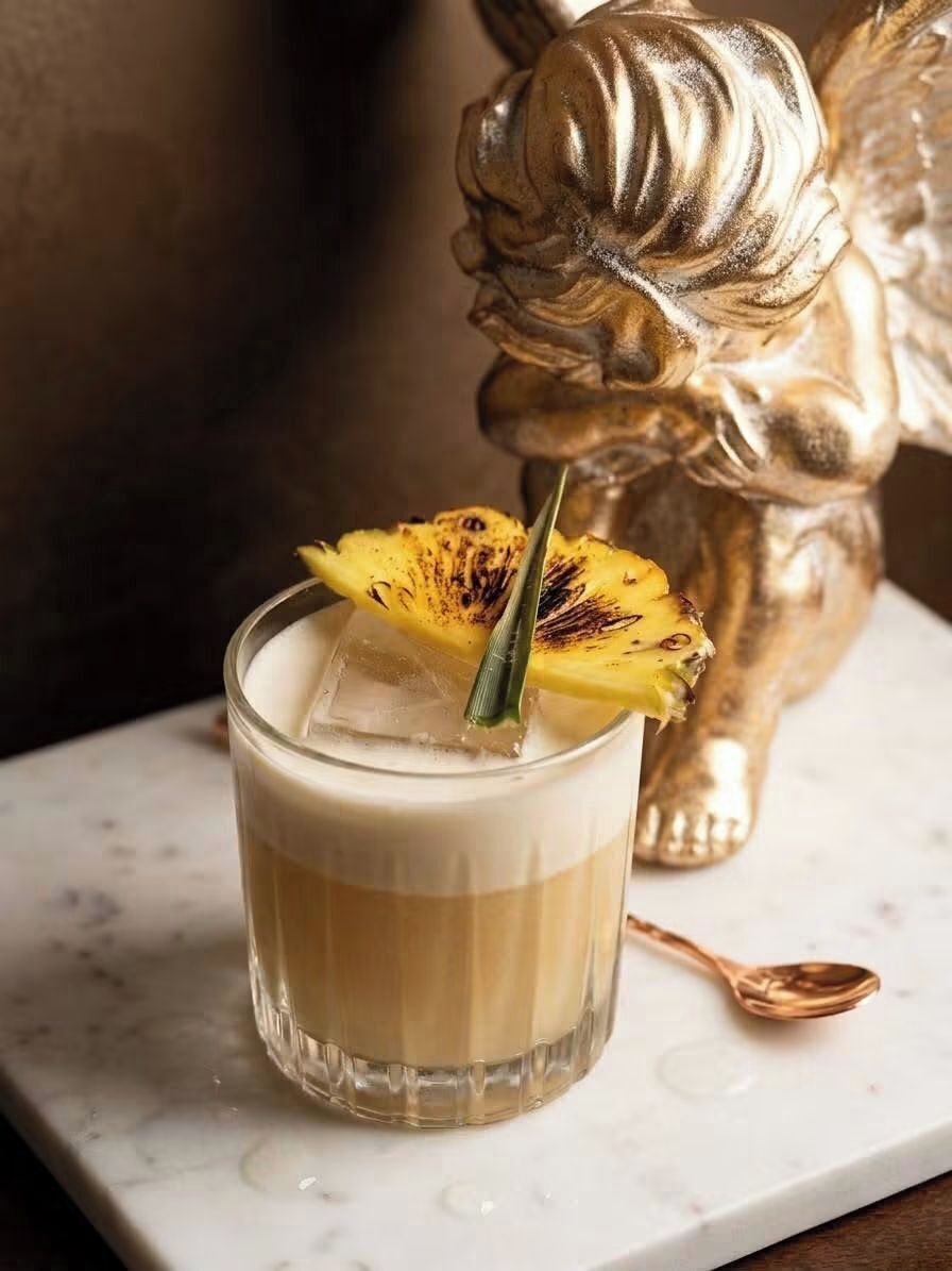 A glass with a layered drink, garnished with a pineapple slice, on a marble surface with a golden statue in the background.
