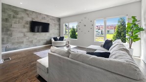 Ein modernes Wohnzimmer mit einer großen Eckcouch, einem weißen Sessel, einem Couchtisch und einem Flachbildfernseher, der an einer strukturierten Wand angebracht ist.