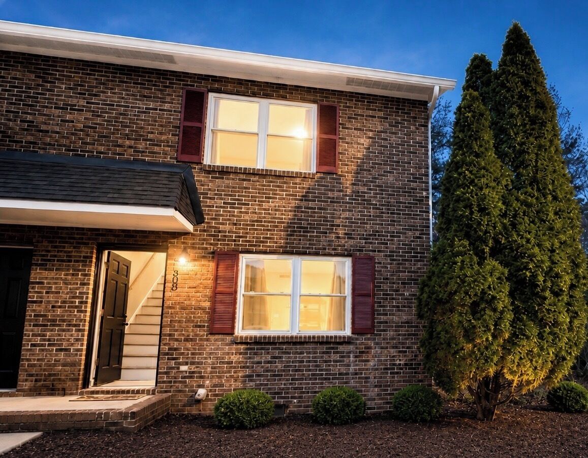 Een twee verdiepingen tellend huis van baksteen, met een duidelijke voordeur, twee ramen met luiken en een verzorgd gazon.