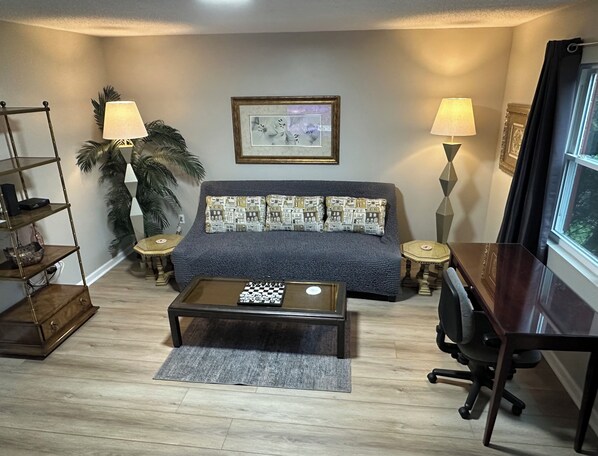 A cozy living room with a sofa, coffee table, and a desk with a chair. There are two lamps, a framed picture on the wall, and a plant.