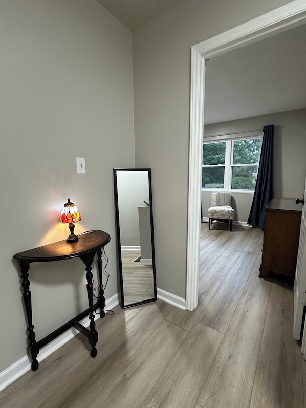 A room with a wooden floor, a table with a lamp, a mirror, and a window with curtains.