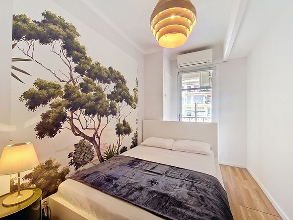A bedroom with a bed, a wall-mounted air conditioner, a window with a view of buildings, and a hanging light fixture.
