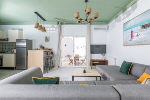 A modern living room with a grey sofa, a wooden coffee table, and a TV. There is a kitchen area with a fridge and a dining table.
