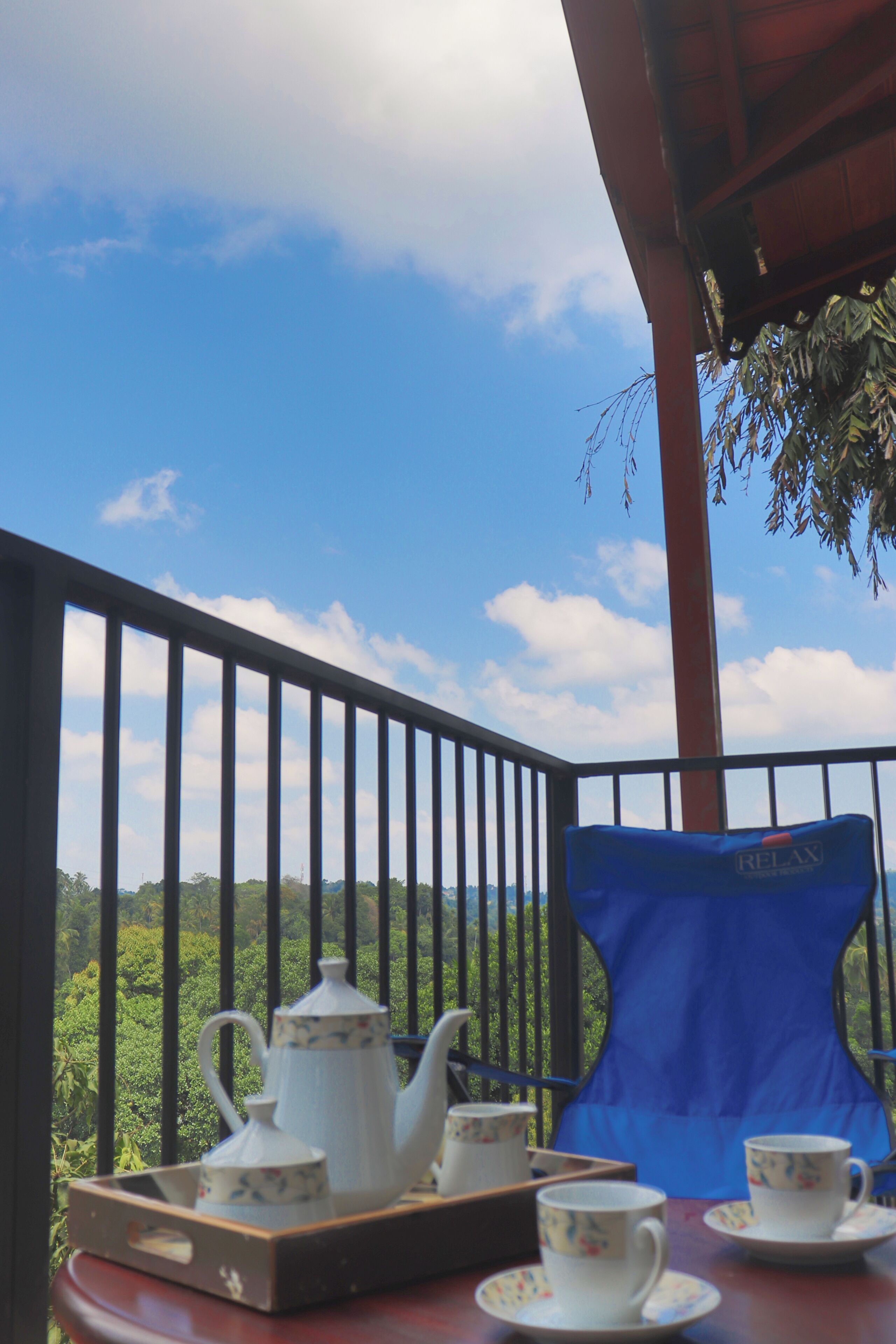 A balcony with a table set for tea, a blue chair, and a view of a lush landscape.
