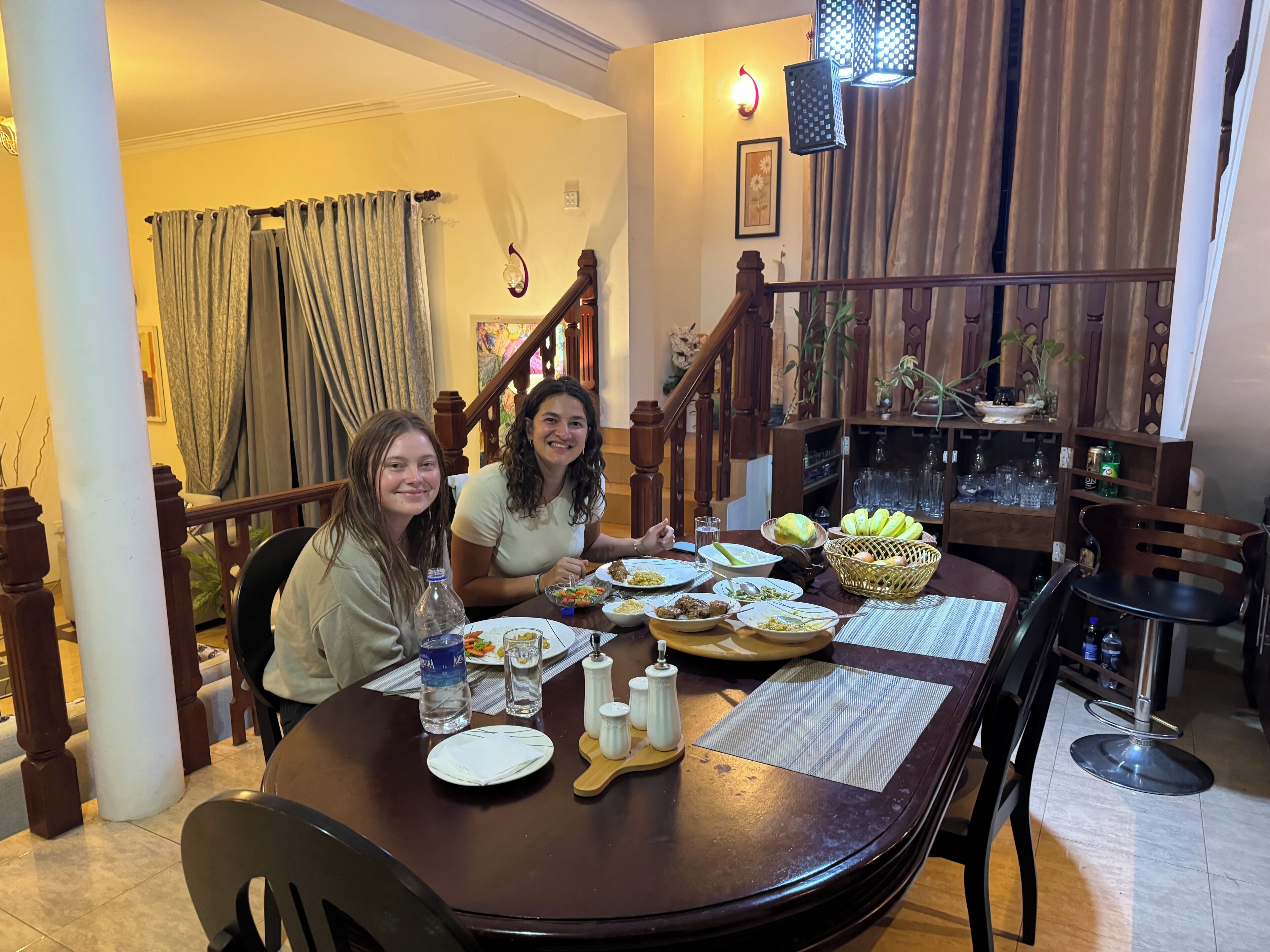 Un comedor con una mesa de madera dispuesta para dos personas, rodeada de sillas. Hay platos con comida, una botella de agua y un salero sobre la mesa. Se ve una escalera con barandillas de madera al fondo.