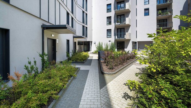Ein modernes Gebäude mit Innenhof, mit gepflastertem Weg und begrünten Flächen mit verschiedenen Pflanzen.