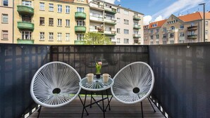Eine Dachterrasse mit Tisch und zwei Stühlen, einem Blumenvasen und Blick auf mehrstöckige Gebäude mit Balkonen.