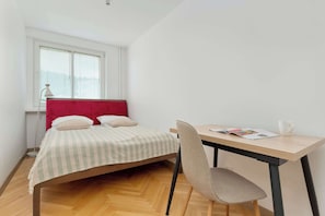 Et soveværelse med seng, skrivebord, stol, et vindue med rullegardiner og en lampe.