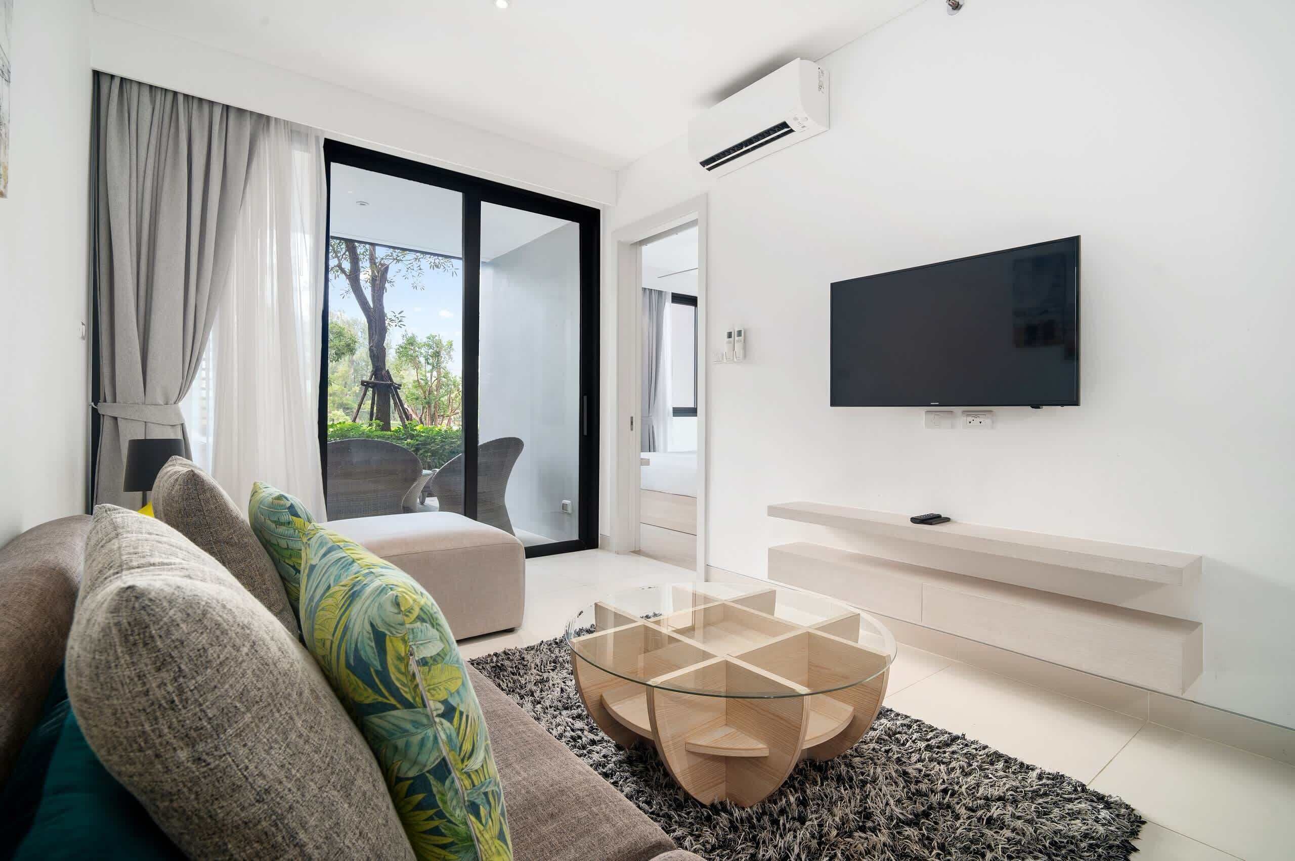 A modern living room with a glass coffee table, a sofa with patterned cushions, a flat-screen TV mounted on the wall, and a sliding glass door leading to an outdoor area.