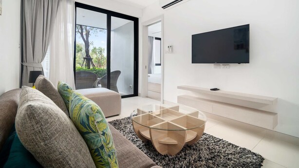 A modern living room with a glass coffee table, a sofa with patterned cushions, a flat-screen TV mounted on the wall, and a sliding glass door leading to an outdoor area. - Cassia Residences, One Bedroom 1103 A1 Ground Floor (Choeng Thale)