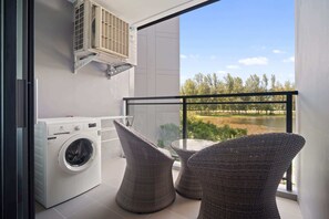 Ein Balkon mit Waschmaschine, Korbstühlen und Blick auf einen See und Bäume.