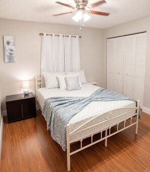 Um quarto com cama, mesa de cabeceira, ventilador de teto, janela com cortinas e armário.