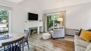 A living room with a fireplace, a television mounted above it, a sofa, a dining table with chairs, and a large window with curtains. - Condo Steps from Lake Pinneo, Fairway Village (Hartford)