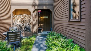 A house with a wooden exterior, a front porch with a bench, a stack of firewood, and a small garden area with plants. - Condo Steps from Lake Pinneo, Fairway Village (Hartford)