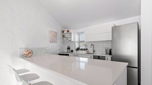 A modern kitchen with white cabinetry, a stainless steel refrigerator, and a large island with bar stools. - Sea Breeze - Orewa Oceanside Apartments (Orewa)