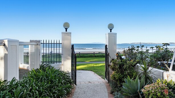 A gated entrance with a gravel path leading to a grassy area and a view of the ocean. - Sea Breeze - Orewa Oceanside Apartments (Orewa)