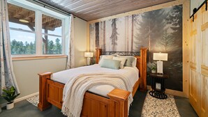 Un dormitorio con una cama grande, cabecera de madera, una ventana con cortinas, un mural temático de bosque y una mesita con lámpara.