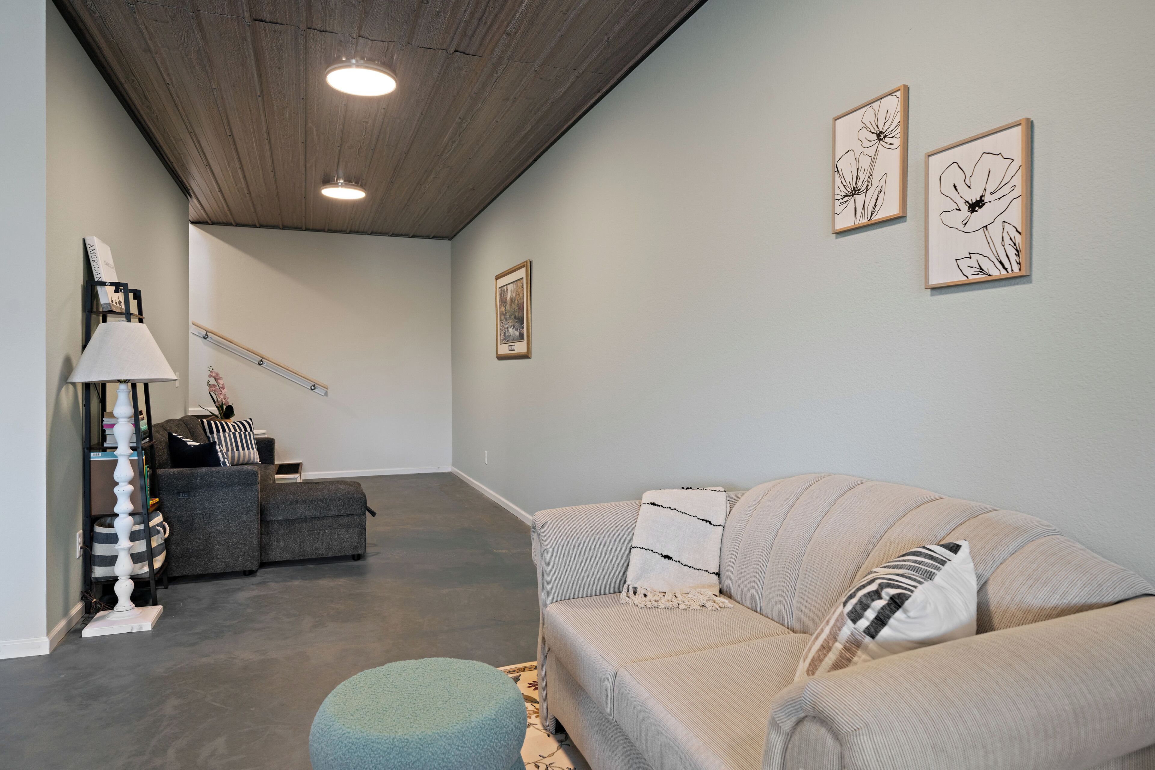 Un salon moderne avec un canapé gris, un pouf bleu et un escalier menant à un autre niveau.