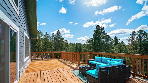 Una terraza de madera con mobiliario exterior, barandilla y vistas a árboles y al cielo.