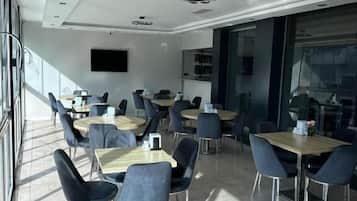 A modern dining area with multiple tables and chairs, large windows allowing natural light, and a flat-screen TV mounted on the wall.