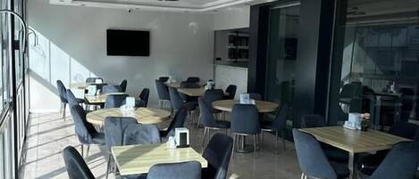 Un comedor moderno con varias mesas y sillas, amplias ventanas que dejan entrar luz natural y un televisor de pantalla plana montado en la pared.
