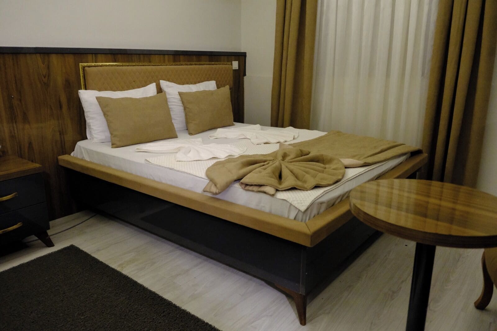 Una cama con cabecera de madera, una mesita de noche, una pequeña mesa redonda y una alfombra oscura.