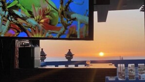 A room with a television displaying a colorful image, a view of the sunset over the sea, and a balcony with a railing. - Your Second Home Awaits – Cozy Stay with Outstanding Service (Agadir)