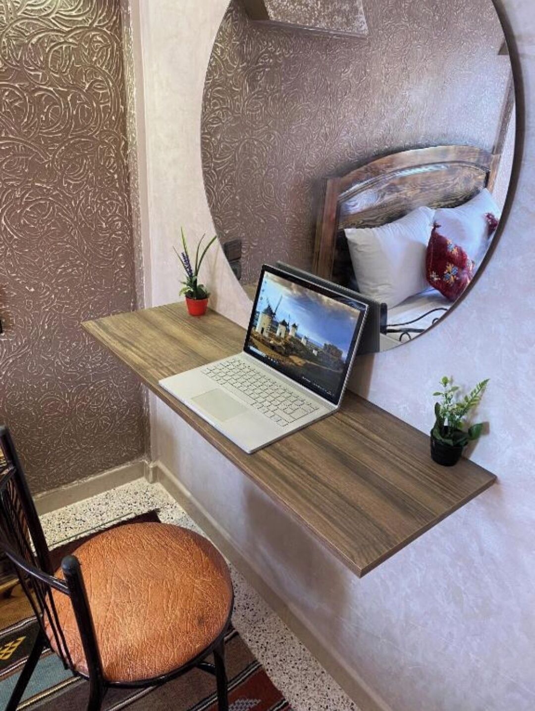 A laptop on a wooden desk with a small potted plant, a chair, and a mirror reflecting a pillow and a red cushion.