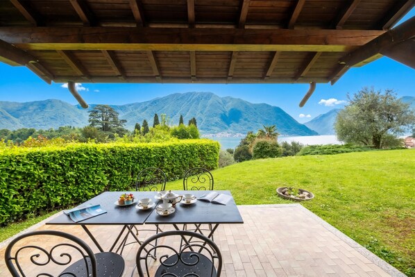 A covered outdoor seating area with a table set for tea, chairs, and a view of mountains and a lake.