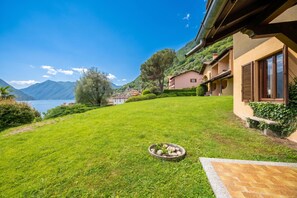 A well-maintained lawn, a stone planter with plants, and a building with a balcony overlooking a lake and mountains.