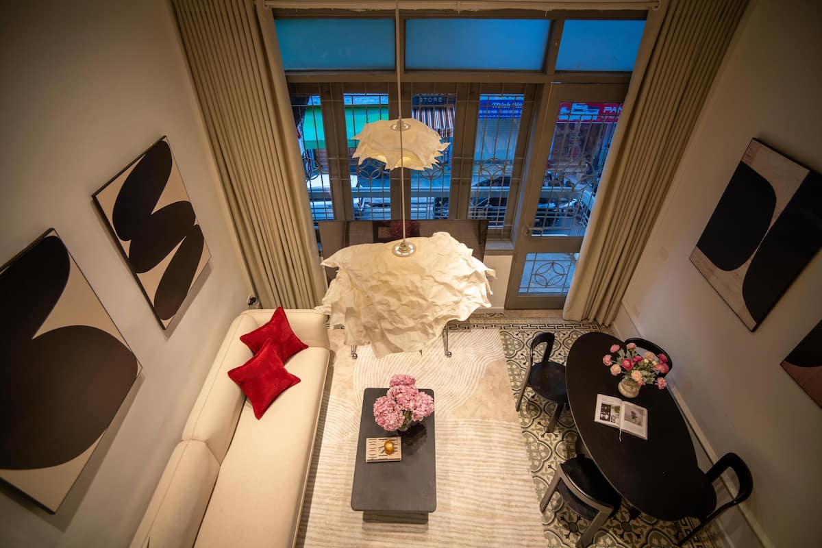 Un salon moderne avec un canapé blanc, un coussin rouge et une table basse ornée de fleurs roses. On aperçoit une table à manger avec des chaises à l’arrière-plan.