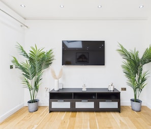 Un soggiorno moderno con una televisione a schermo piatto montata a parete, un mobile TV in legno scuro con cassetti e due piante in vaso su entrambi i lati.