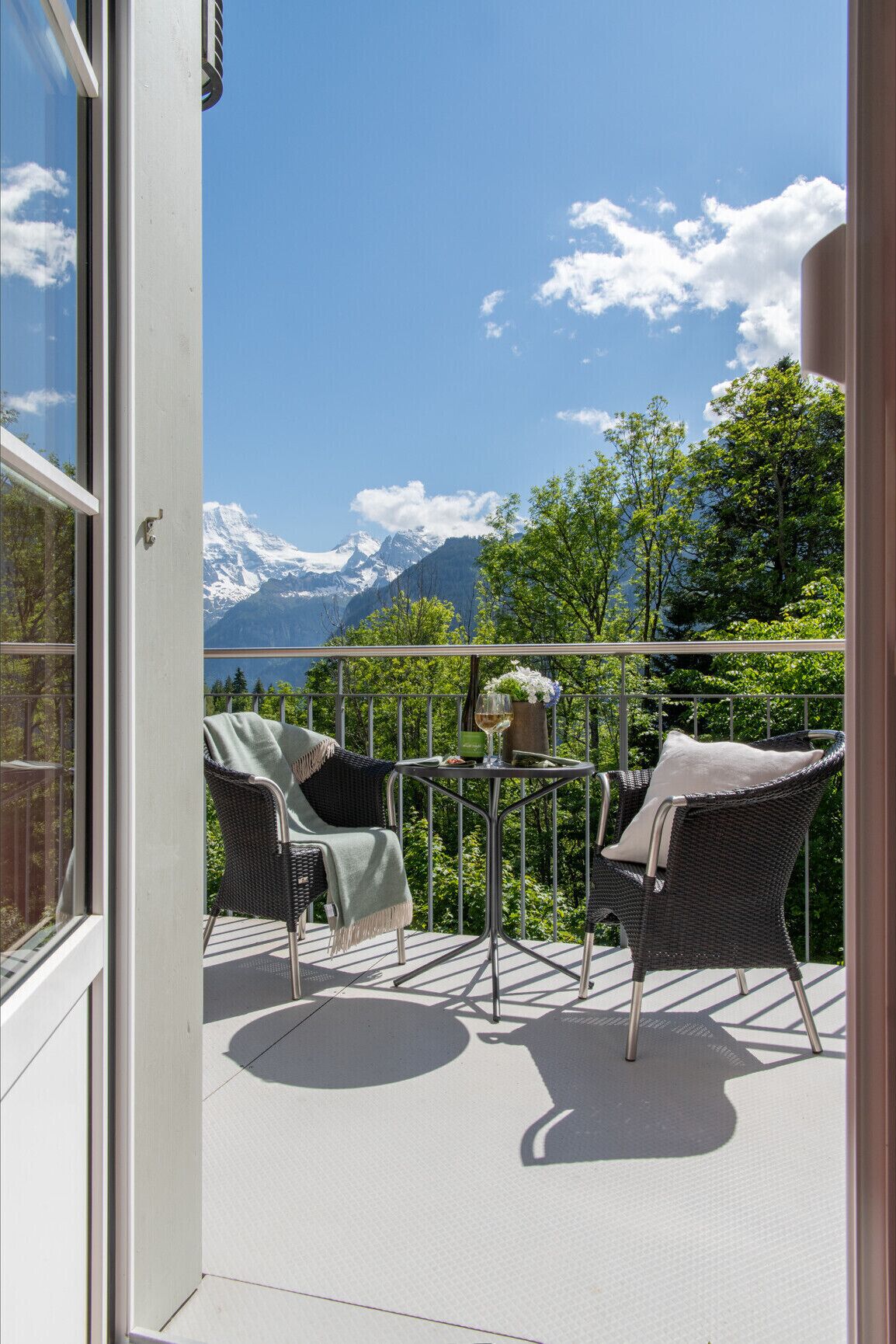 Een balkon met rieten stoelen en een tafel, uitkijkend op een berglandschap.