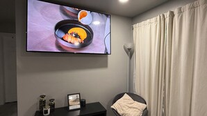 Una sala moderna con un televisor de pantalla plana montado en la pared, un mueble de entretenimiento negro con un cuadro enmarcado, una silla negra con una manta blanca y una lámpara de pie.