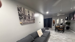 Una sala de estar moderna con un sofá gris, una mesa de comedor con sillas, un ventilador de techo y un cuadro con una vista urbana en la pared.