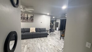 Una sala de estar con un sofá gris, una mesa de comedor con sillas, un ventilador de techo y un cuadro en la pared.