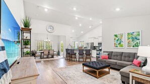 A spacious living room with a large TV, a grey sofa, and a dining area with a kitchen in the background. - 3BR Legends Golf Home | Bunk Room + Backyard (Myrtle Beach)