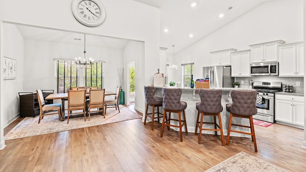 A spacious kitchen with white cabinets, a stainless steel refrigerator, and a microwave. There is a dining area with a wooden table and chairs, and a chandelier hanging above. - 3BR Legends Golf Home | Bunk Room + Backyard (Myrtle Beach)
