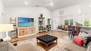 A spacious living room with a large flat-screen TV, wooden cabinets, a black ottoman, and a dining area with bar stools. - 3BR Legends Golf Home | Bunk Room + Backyard (Myrtle Beach)