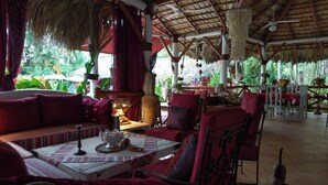 An outdoor seating area with red upholstered chairs and a table, surrounded by lush greenery and a thatched roof structure. - Belle propriété de style Caraïbes, confortable chambre d'hôtes, piscine privée (Las Terrenas)